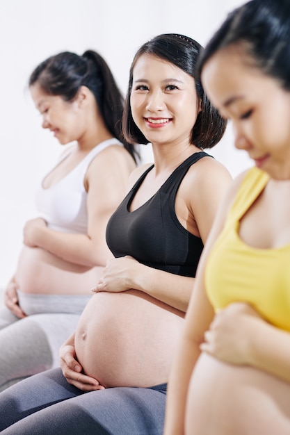 Fröhliche hübsche junge vietnamesische Frau, die ihren Bauch berührt und massiert, wenn sie Schwangerschaftsklasse besucht