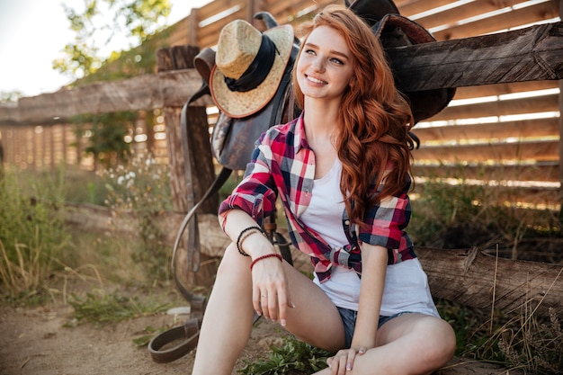 Fröhliche hübsche rothaarige junge Frau Cowgirl, die draußen sitzt und lächelt