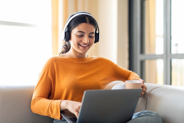 Fröhliche indische Frau genießt Musik mit Kopfhörern, während sie an ihrem Laptop arbeitet