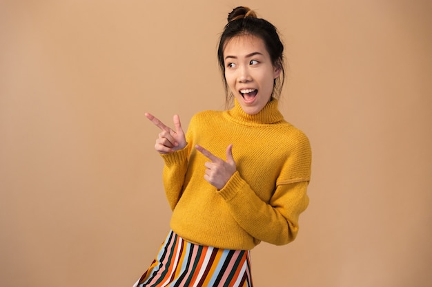 Fröhliche japanische Frau mit Pullover lächelt und zeigt mit den Fingern beiseite auf Kopienraum isoliert über beige Wand im Studio