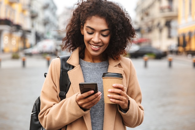 Fröhliche junge afrikanische Frau, die Mantel trägt, der draußen geht, Kaffeetasse zum Mitnehmen hält, mit Handy