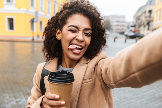 Fröhliche junge afrikanische Frau, die Mantel trägt, der draußen geht, Kaffeetasse zum Mitnehmen hält und ein Selfie nimmt