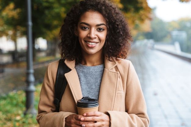 Fröhliche junge afrikanische Frau, die Mantel trägt, der draußen geht und Kaffeetasse zum Mitnehmen hält