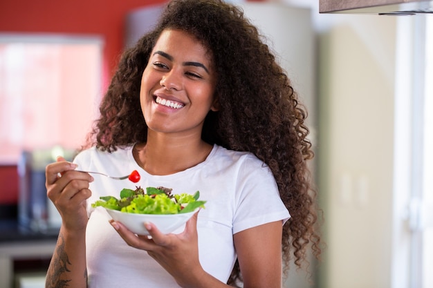 fröhliche junge afroamerikanische Frau, die Gemüsesalat in der Hauptküche isst