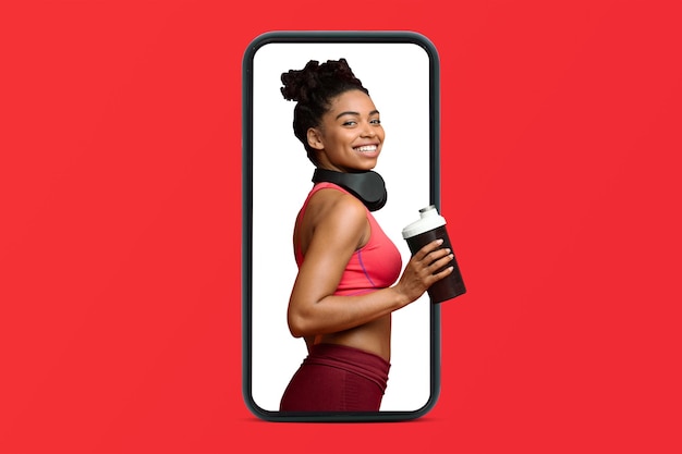 Fröhliche junge afroamerikanische schlanke Dame in Sportbekleidung und Kopfhörern mit Cocktail auf dem Smartphone