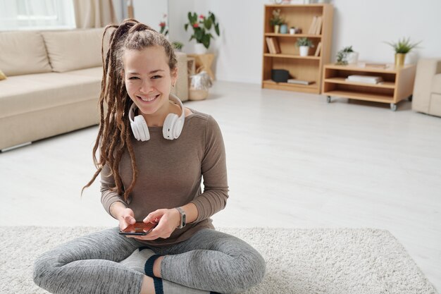 Fröhliche junge aktive Frau mit Kopfhörern unter Verwendung des Smartphones beim Üben des Yoga in der häuslichen Umgebung