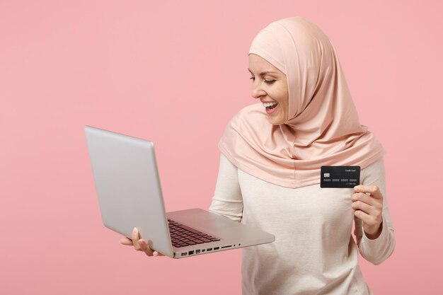 Fröhliche junge arabische muslimische Frau in Hijab-leichter Kleidung posiert isoliert auf rosa Hintergrund. Menschen religiösen Islam Lifestyle-Konzept. Mock-up-Kopienbereich. Halten Sie Laptop-PC-Computer, Kreditkarte.