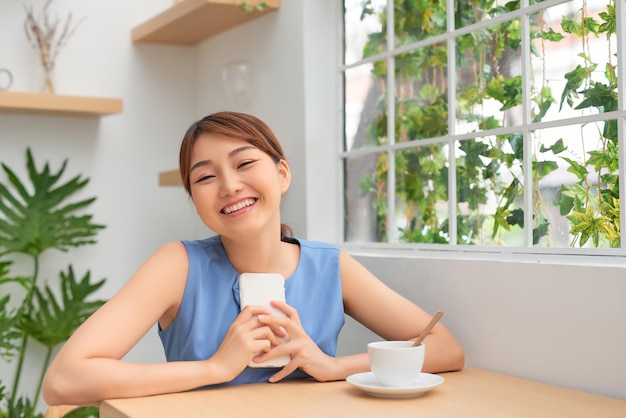 Fröhliche junge Asiatin, die lächelt, während sie telefoniert und Kaffee trinkt