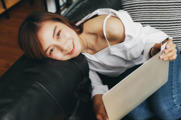 Fröhliche junge Asiatin mit Kreditkarte und Laptop, die zu Hause auf dem Sofa sitzt