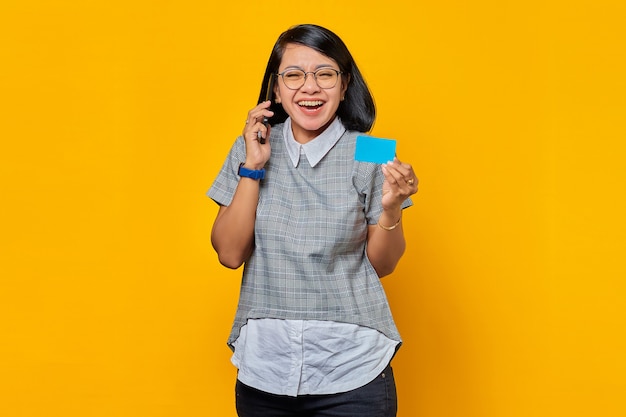 Fröhliche junge asiatische Frau, die auf dem Smartphone spricht und Kreditkarte auf gelbem Hintergrund zeigt?