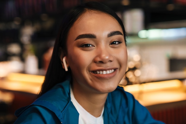 fröhliche junge asiatische frau, die drahtlose kopfhörer verwendet und lächelt, während sie im café sitzt