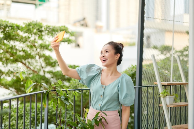 Fröhliche junge asiatische Frau, die einen Videoanruf mit ihrem Telefon beim Stehen o Balkon hat