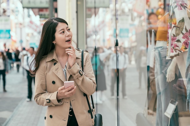 fröhliche junge asiatische frau, die schaufenster lächelt und schaut. glückliche bürodame genießt die freizeit nach der arbeit in dotonbori. mode weibliche shopaholic halten handy und schockierte glamouröse kleidung im laden