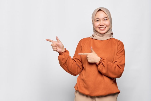 Fröhliche junge asiatische Frau im orangefarbenen Hemd, die mit dem Finger auf den Kopierraum zeigt, der über weißem Hintergrund isoliert ist