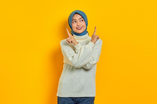Fröhliche junge asiatische Frau im weißen Pullover, die mit dem Finger nach oben auf den Kopierbereich zeigt, der über gelbem Hintergrund isoliert ist