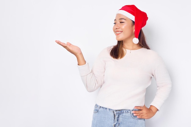 Fröhliche junge asiatische Frau in einer Weihnachtsmütze, die Kopienraum auf der Handfläche isoliert über weißem Hintergrund präsentiert Feier Weihnachtsfeiertag und Neujahrskonzept