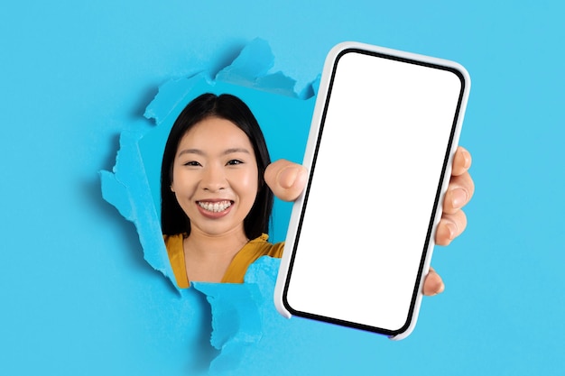 Fröhliche junge asiatische Frau lächelt durch ein zerrissenes blaues Papier und präsentiert ein Smartphone mit einem leeren