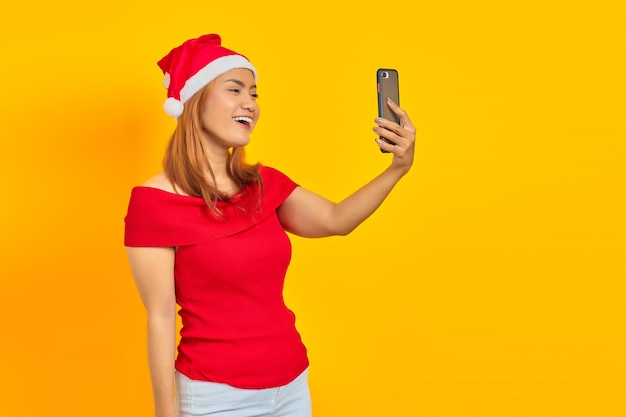 Fröhliche junge asiatische Frau mit Weihnachtsmütze macht Videoanruf auf gelbem Hintergrund