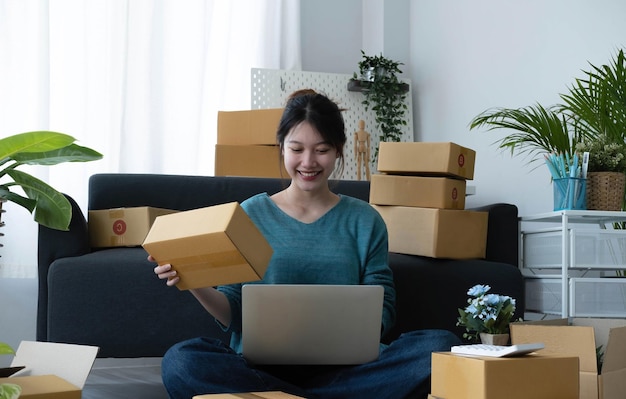 Fröhliche junge asiatische Frau Startup Kleinunternehmen freiberuflich tätig, die Paketbox und Computer-Laptop hält und auf dem Boden sitzt Online-Marketing-Verpackungsbox-Lieferkonzept
