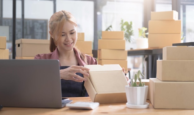 Fröhliche junge asiatische Frau Startup Kleinunternehmen freiberuflich tätig, die Paketbox und Computer-Laptop hält und auf einem Stuhl sitzt Online-Marketing-Verpackungsbox-Lieferkonzept