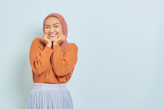 Fröhliche junge asiatische Muslimin in braunem Pullover und Handflächen am Kinn isoliert auf weißem Hintergrund Muslimisches Lifestyle-Konzept