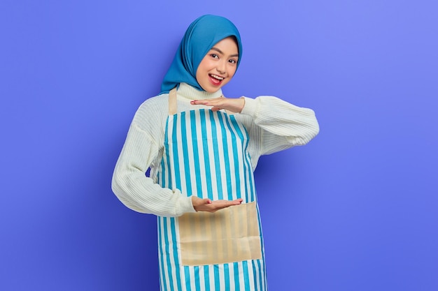 Fröhliche junge asiatische Muslimin in Hijab und gestreifter Schürze, die Produkte mit Palmen zeigt, die Platz für bestimmte Rabatte auf violettem Hintergrund zeigen Menschen Hausfrau muslimischen Lifestyle-Konzept