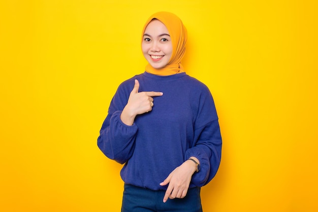 Fröhliche junge asiatische Muslimin in lässigem Pullover, die stolz isoliert auf gelbem Hintergrund auf sich selbst zeigt
