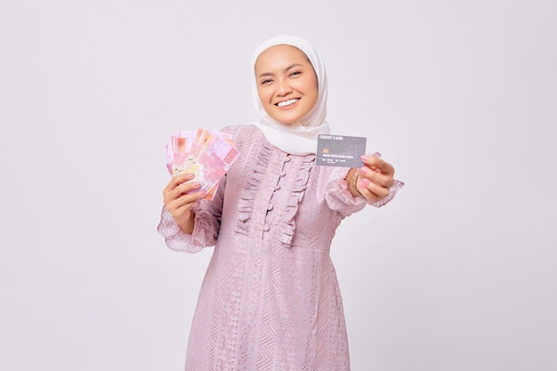 Fröhliche junge asiatische Muslimin mit Hijab und lila Kleid mit Kreditkarte und Geldscheinen isoliert auf weißem Studiohintergrund