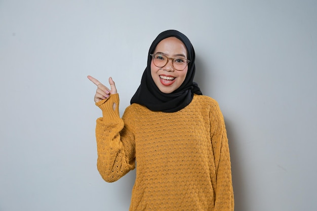 Fröhliche junge asiatische Muslimin mit orangefarbenem Pullover und Brille zeigt auf den leeren Bereich daneben
