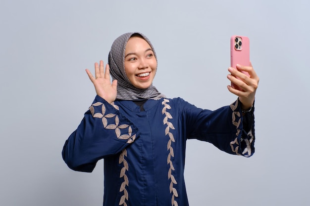 Fröhliche junge asiatische Muslimin mit Smartphone, die einen Videoanruf tätigt oder ein Selfie isoliert auf weißem Hintergrund macht