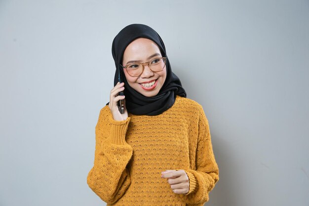 Fröhliche junge asiatische Muslimin trägt orangefarbenen Pullover und Brille, während sie ihr Mobiltelefon benutzt