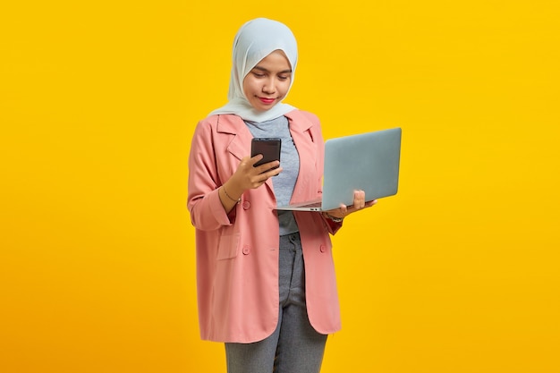 Fröhliche junge attraktive Frau mit Laptop und Handy im Stehen isoliert auf gelbem Hintergrund