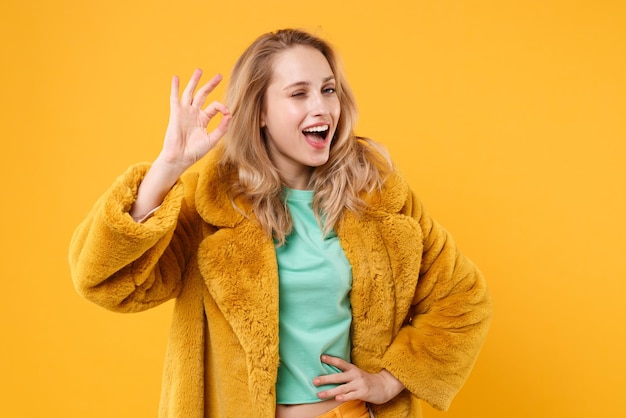 Fröhliche junge blonde Frau Mädchen im gelben Pelzmantel posiert isoliert auf orangefarbenem Hintergrund Studioportrait. Menschen aufrichtige Emotionen Lifestyle-Konzept. Mock-up-Kopienbereich. Blinkt, zeigt OK-Geste.