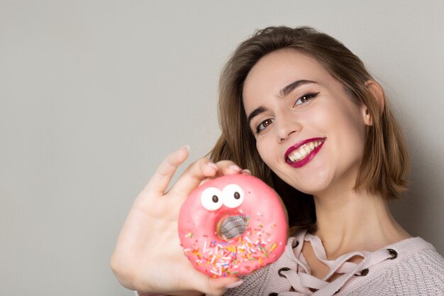 Fröhliche junge brünette Frau mit süßem Donut mit rosa Glasur posiert in der Nähe von grauem Hintergrund