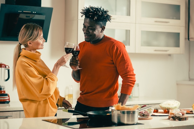 Fröhliche junge Dame, die zu ihrem glücklichen Freund lächelt, während sie mit ihm in der Küche steht und köstlichen Rotwein genießt