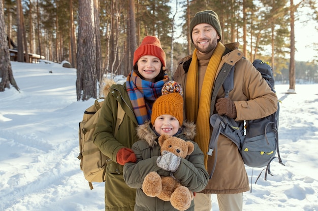 Fröhliche junge dreiköpfige Familie in warmer Winterkleidung, die vor der Kamera gegen die natürliche Umgebung steht und Sie draußen ansieht