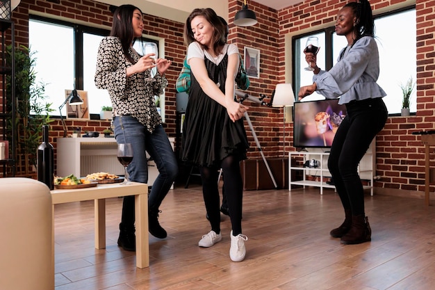 Fröhliche junge enge Freunde tanzen zusammen im Wohnzimmer, während sie Geburtstag feiern. Fröhliche Menschen im Wohnzimmer zu Hause genießen eine Weinparty, während sie trinken und Spaß haben.