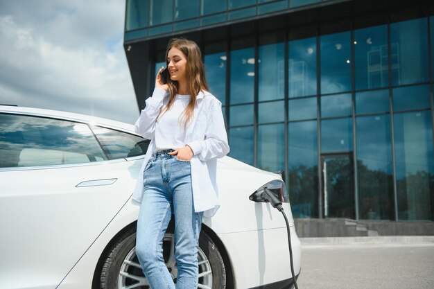 Fröhliche junge erwachsene Frau, die breit lächelt und wegschaut, um die Autobatterie von einer kleinen öffentlichen Station aufzuladen, die in der Nähe eines Elektroautos steht