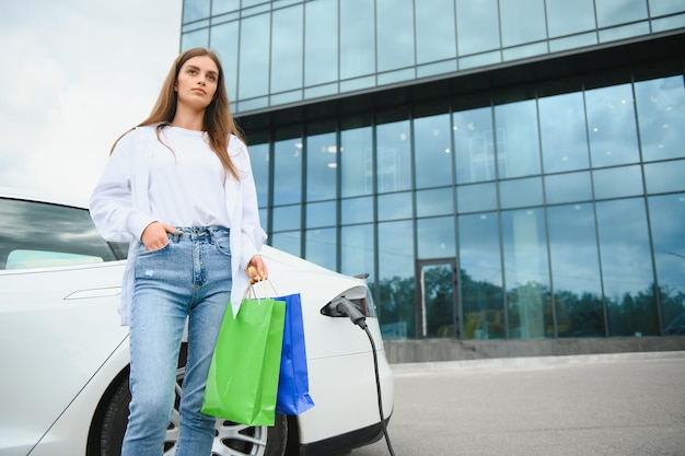 Fröhliche junge erwachsene Frau, die breit lächelt und wegschaut, um die Autobatterie von einer kleinen öffentlichen Station aufzuladen, die in der Nähe eines Elektroautos steht