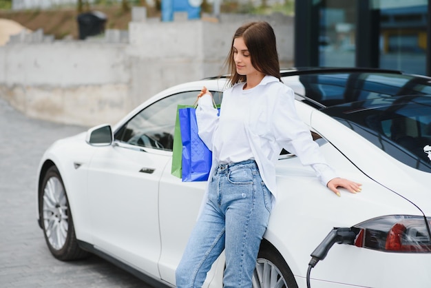 Fröhliche junge erwachsene Frau, die breit lächelt und wegschaut, um die Autobatterie von einer kleinen öffentlichen Station aufzuladen, die in der Nähe eines Elektroautos steht