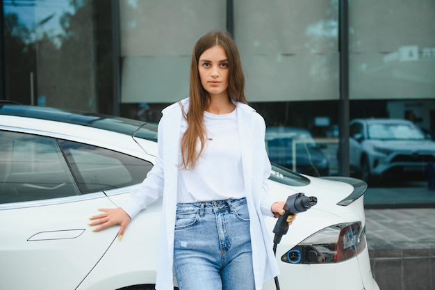 Fröhliche junge erwachsene Frau, die breit lächelt und wegschaut, um die Autobatterie von einer kleinen öffentlichen Station aufzuladen, die in der Nähe eines Elektroautos steht