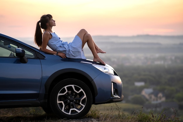 Fröhliche junge Fahrerin in blauem Kleid, die einen warmen Sommerabend genießt, der auf ihrer Motorhaube liegt Reise- und Urlaubskonzept