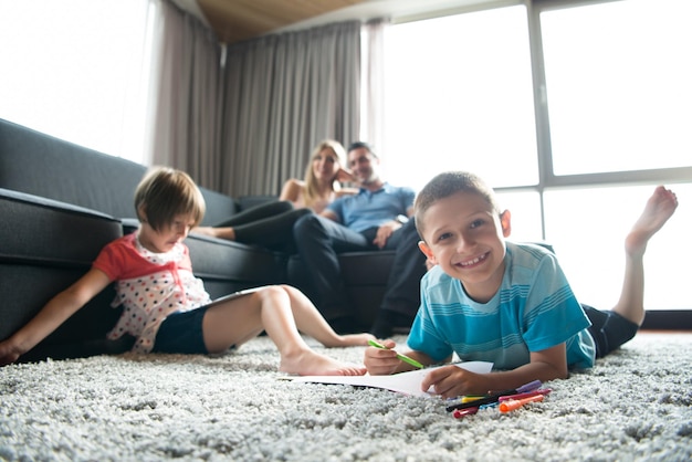 Fröhliche junge Familie, die zu Hause auf dem Boden mit einem Tablet und einem Zeichenset für Kinder zusammen spielt