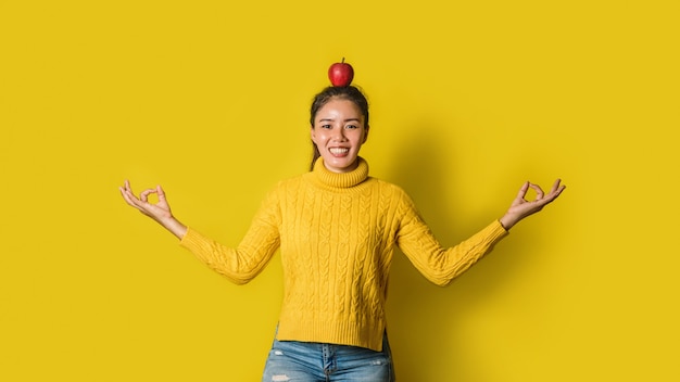 Fröhliche junge Frau auf gelbem Hintergrund im Studio. Ein Mädchen mit einem Apfel, der beim Yoga auf dem Kopf ruht. Das Konzept der Bewegung für eine gute Gesundheit. Gesundheitsliebhaber