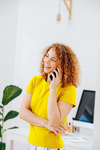 Fröhliche junge Frau, die an ihrem Telefon spricht und vor ihrem Arbeitsplatz steht