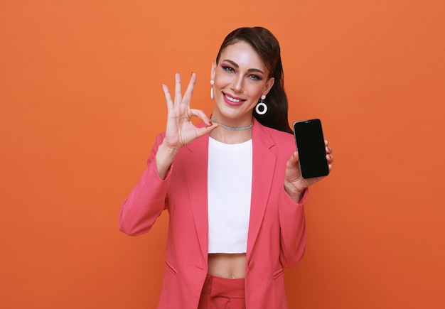 Fröhliche junge Frau, die auf dem Handy mit leerem Bildschirm und der Handbewegung OK auf orangefarbenem Hintergrund zeigt