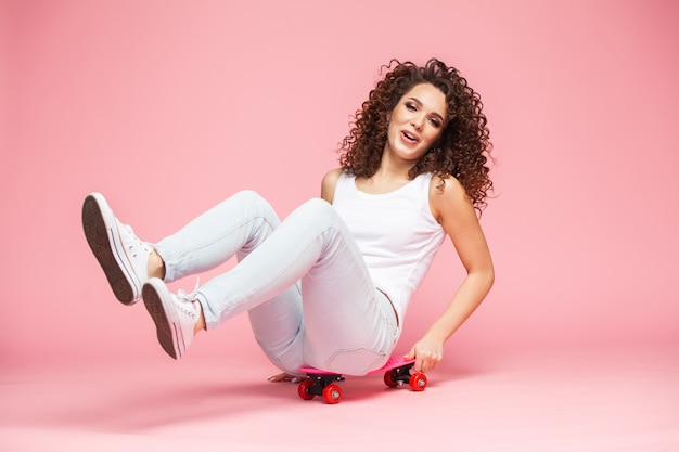 Fröhliche junge Frau, die auf Skateboard sitzt und Spaß über Rosa hat.