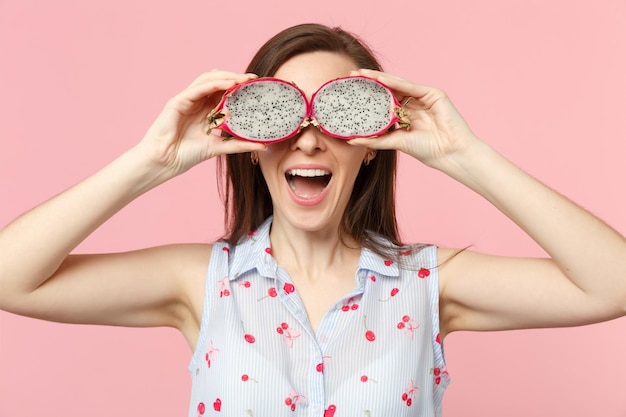 Fröhliche junge Frau, die die Augen mit Hälften frischer, reifer Pitahaya-Drachenfrüchte bedeckt, isoliert auf rosa Pastellwandhintergrund. Menschen lebendiger Lebensstil entspannen Urlaubskonzept. Mock-up-Kopienbereich.