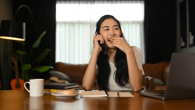 Fröhliche junge Frau, die ein angenehmes Telefongespräch mit ihrer Freundin führt, während sie im gemütlichen Heimbüro sitzt