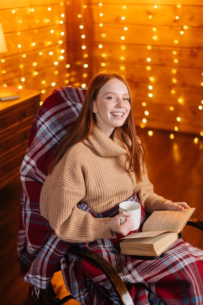 Fröhliche junge Frau, die eine Tasse mit heißem Getränk hält und ein Buch liest, während sie am Schaukelsessel sitzt